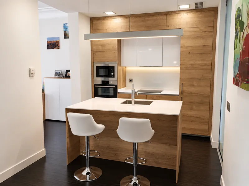 Cocina con isla y taburetes blancos, frente de madera y columna de hornos