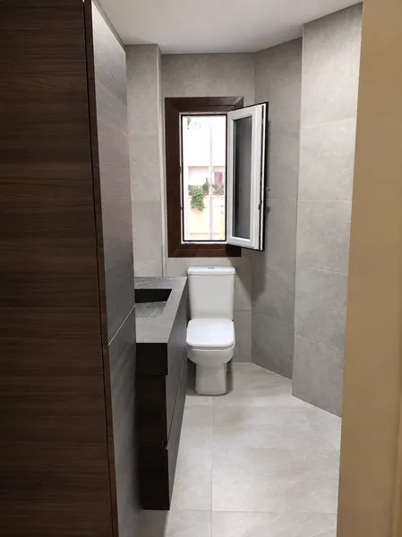 Baño estrecho con revestimiento gris, mueble de madera oscura y ventana al fondo