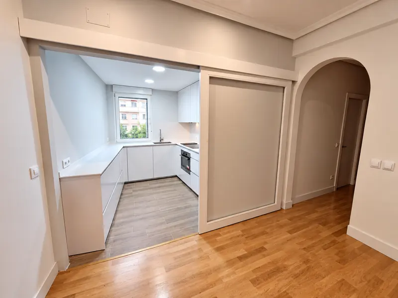 Pasillo reformado con arco y vista a la cocina blanca, con suelo de madera